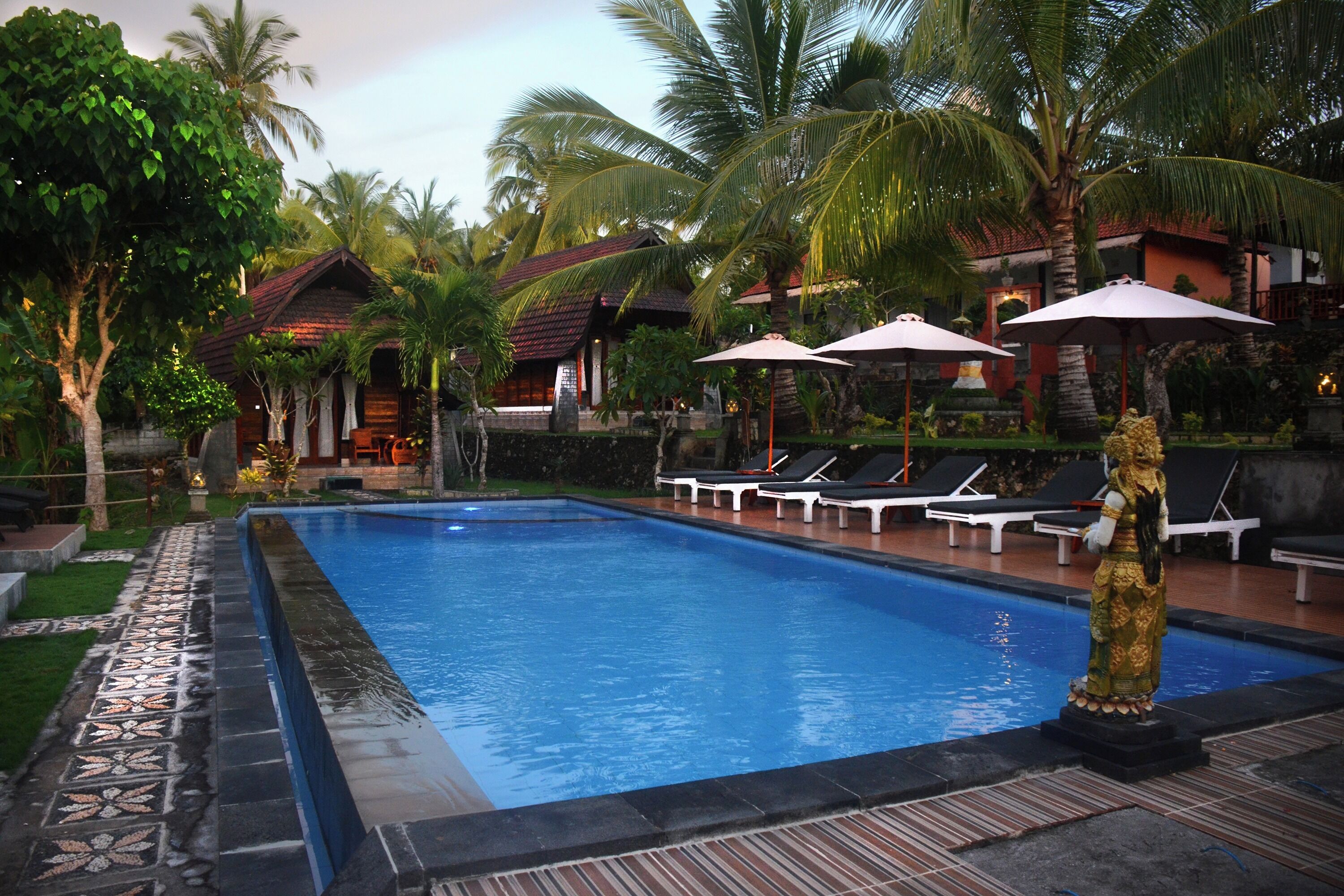 A swimming pool with lounge chairs and umbrellas, surrounded by palm trees and a building with a tiled roof.