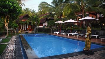 A swimming pool with lounge chairs and umbrellas, surrounded by palm trees and a building with a tiled roof.