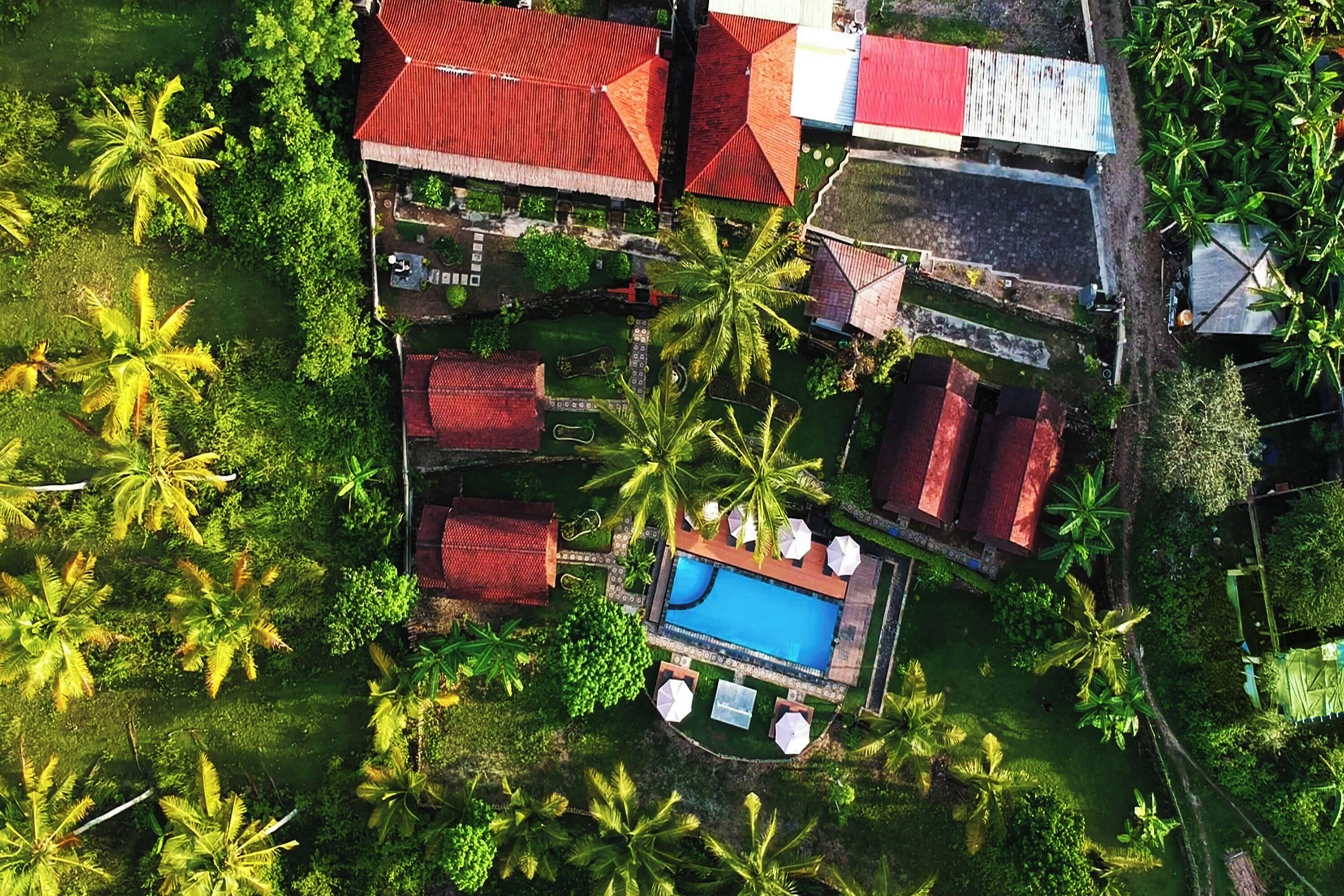 An aerial view of a resort with a swimming pool, palm trees, and red-roofed buildings.