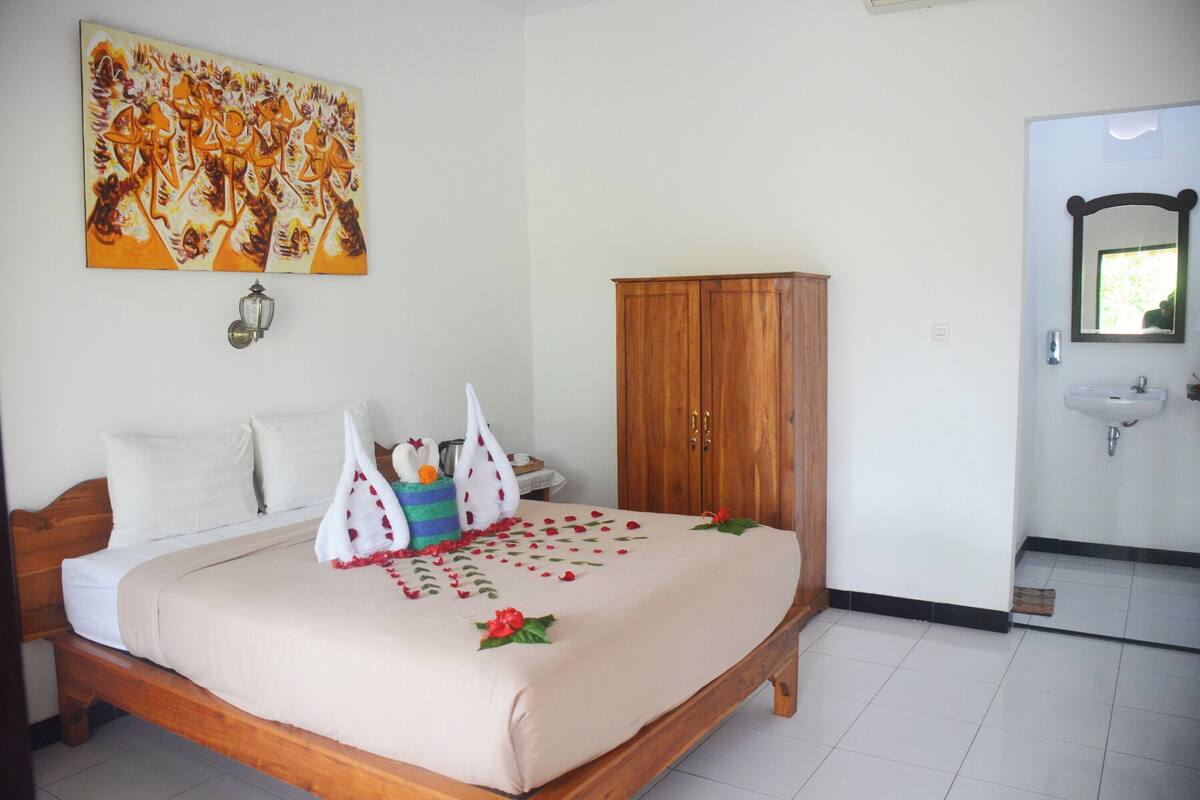 A hotel room with a bed, a wooden wardrobe, a bathroom with a sink and mirror, and a wall with a painting.