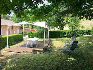 Un patio con tavolo e sedie sotto un tetto bianco, circondato da un giardino curato e un siepe.