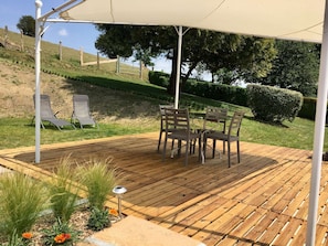 Un terrazzo in legno con tavolo e sedie, riparato da un tetto e circondato da piante e un recinto.