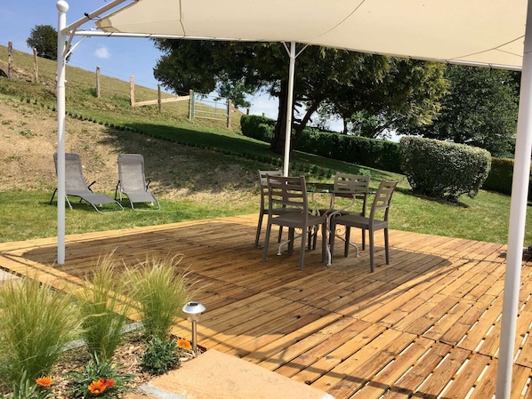 A wooden deck with a table and chairs under a canopy, surrounded by greenery and a fence.