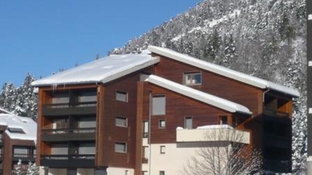 Un immeuble à plusieurs étages, doté d’un toit en pente, vraisemblablement un hôtel ou un complexe touristique, entouré d’arbres couverts de neige et avec une chaîne de montagnes en arrière-plan.