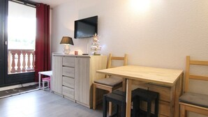Une petite pièce avec une table en bois, deux chaises, un téléviseur fixé au mur, un petit meuble et une fenêtre avec des rideaux rouges.