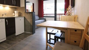 Une cuisine compacte avec un coin repas, un petit réfrigérateur, une machine à café et une vue sur l’extérieur grâce à une fenêtre ornée de rideaux rouges.