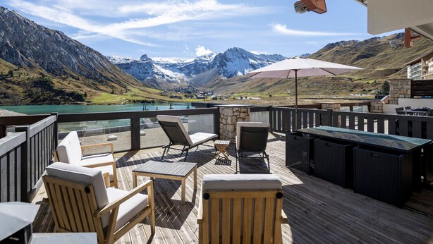 Una terraza de madera con asientos al aire libre, una mesa y un gran paraguas, con vistas a un paisaje montañoso con un lago y picos nevados.