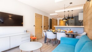 Una sala moderna con un sofá azul, un comedor con sillas, una mesa blanca y una cocina con mostrador y luces colgantes.