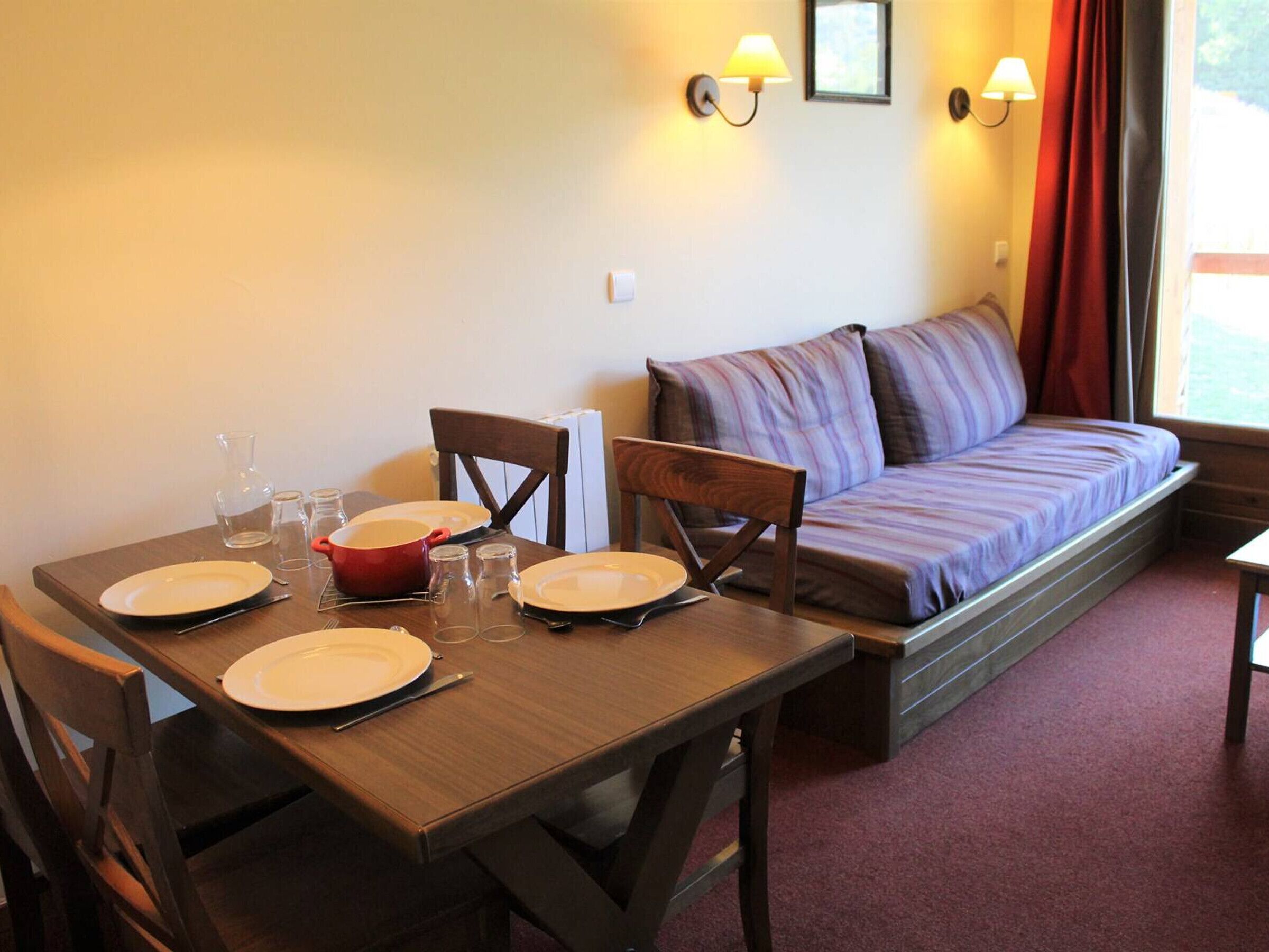 A dining area with a wooden table set for four, a red pot, and two wall-mounted lamps.