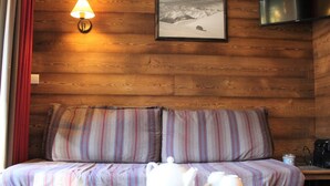 Een gezellig cabin interieur met een houten wand, een bank met gestreepte kussens, een tafel met theepot en kopjes, een lamp, en een lijst met een besneeerd landschap.
