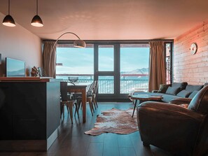 A modern living room with a large window, a sofa, a dining table with chairs, and a small kitchen area.