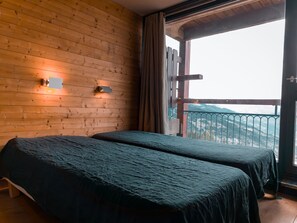 A room with wooden walls, two beds with dark bedding, a window with a view of a mountain, and a wall-mounted light fixture.