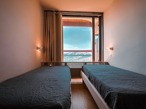 Une chambre avec deux lits, une fenêtre avec des rideaux, un balcon et un luminaire fixé au mur.