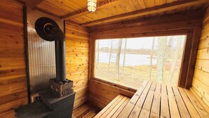 A cabin interior with a wood-burning stove, a window with a view of a lake, and wooden walls.