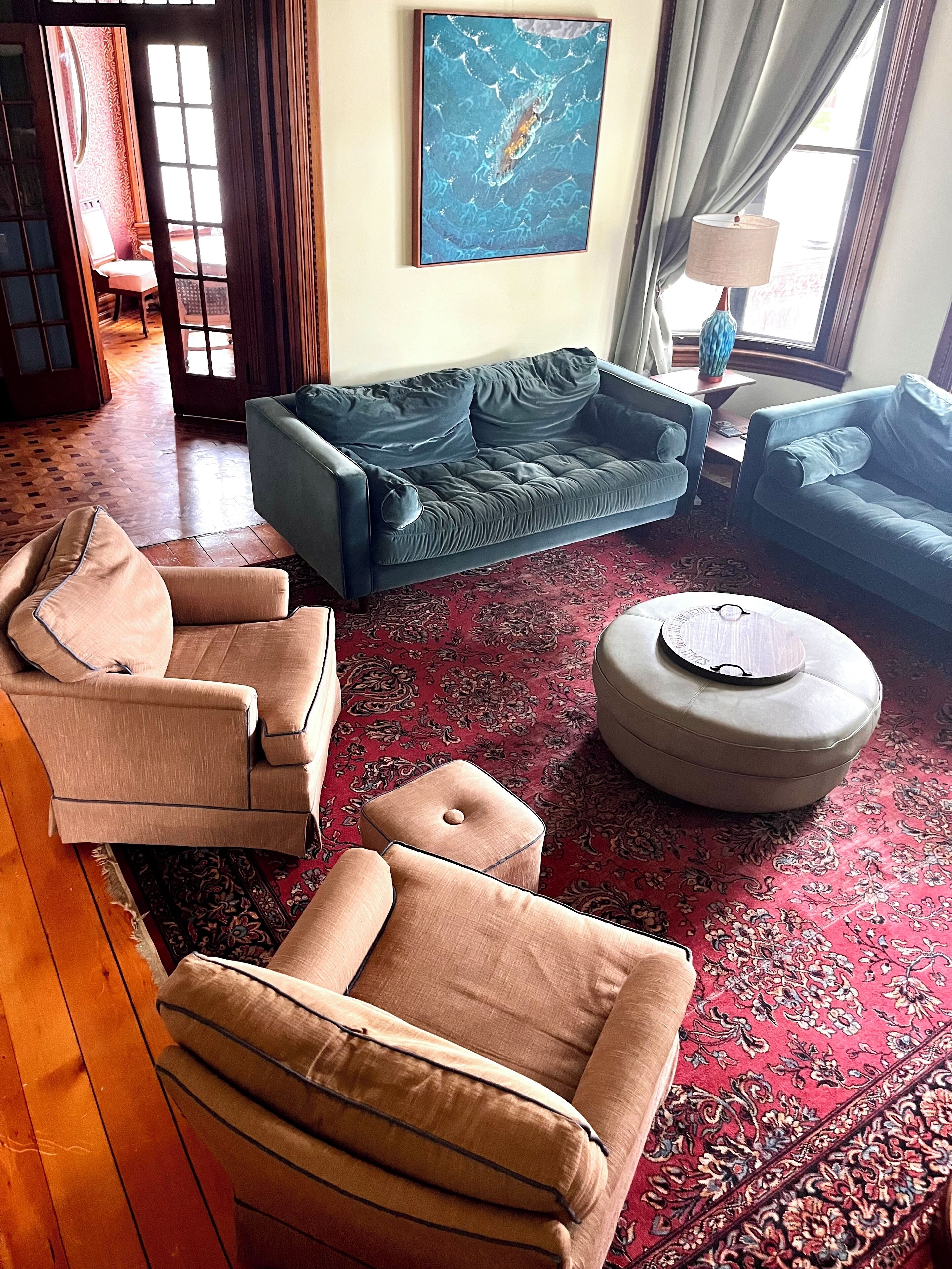Una sala de estar con un sofá azul, sillones color beige, un reposapiés redondo y un tapete rojo estampado.
