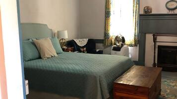 A bedroom with a bed, a wooden chest, a nightstand, a lamp, a clock, a fan, and a fireplace.