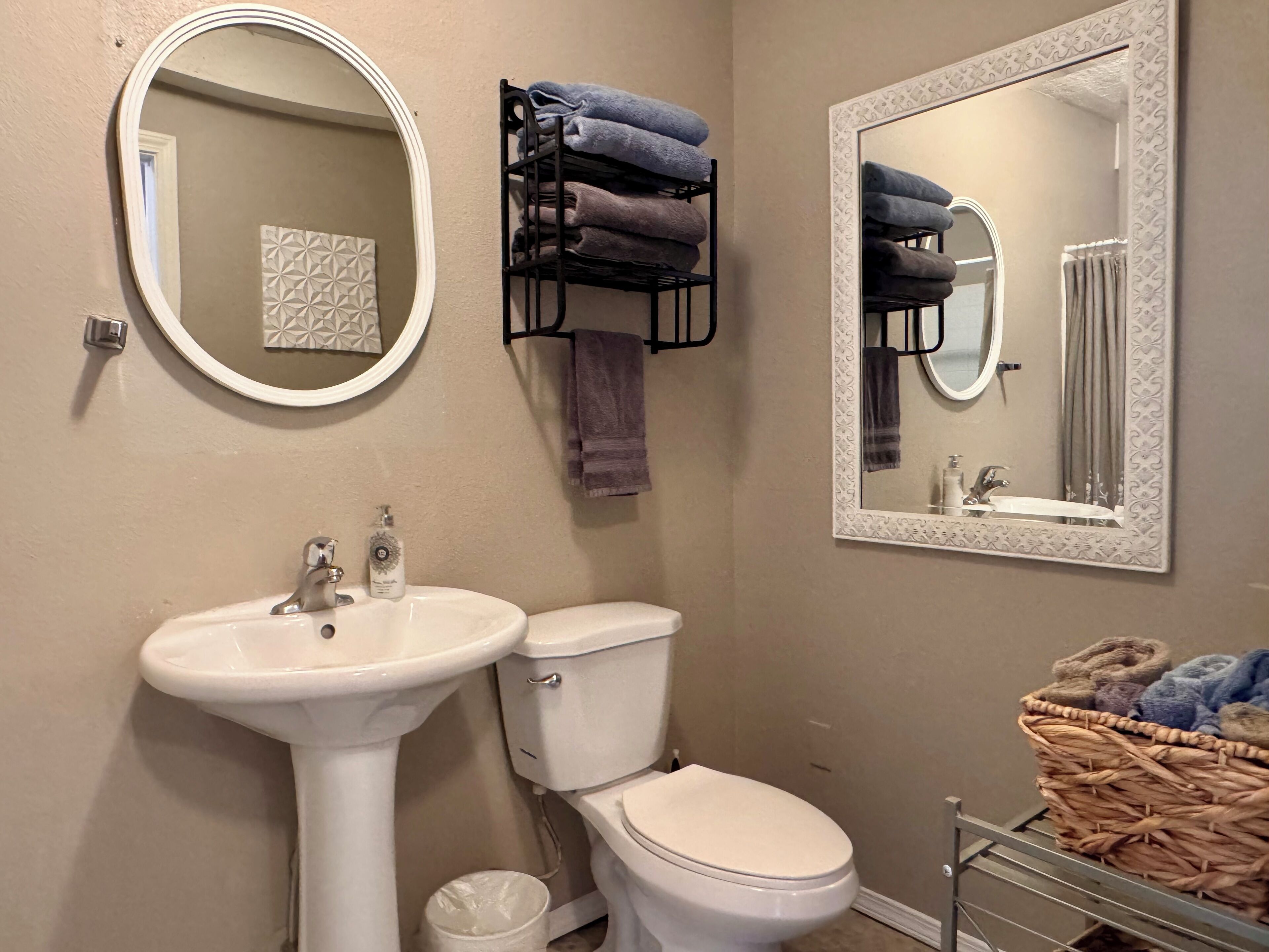 A bathroom with a white sink, toilet, and two mirrors with decorative frames. There are towels stacked on a rack and a basket with towels.