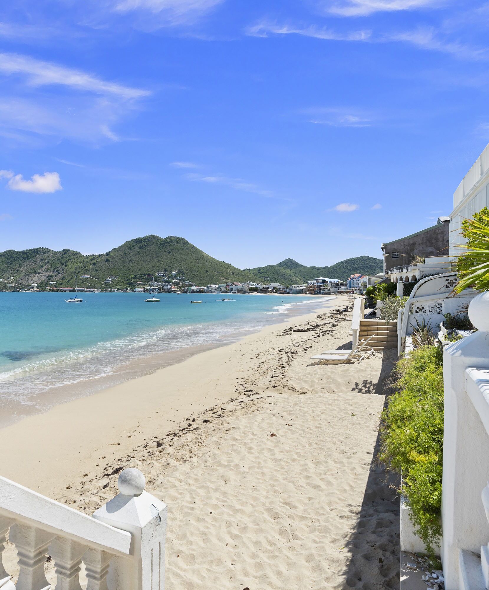 A beach with a sandy path, a white railing, and a view of the sea and hills in the background.