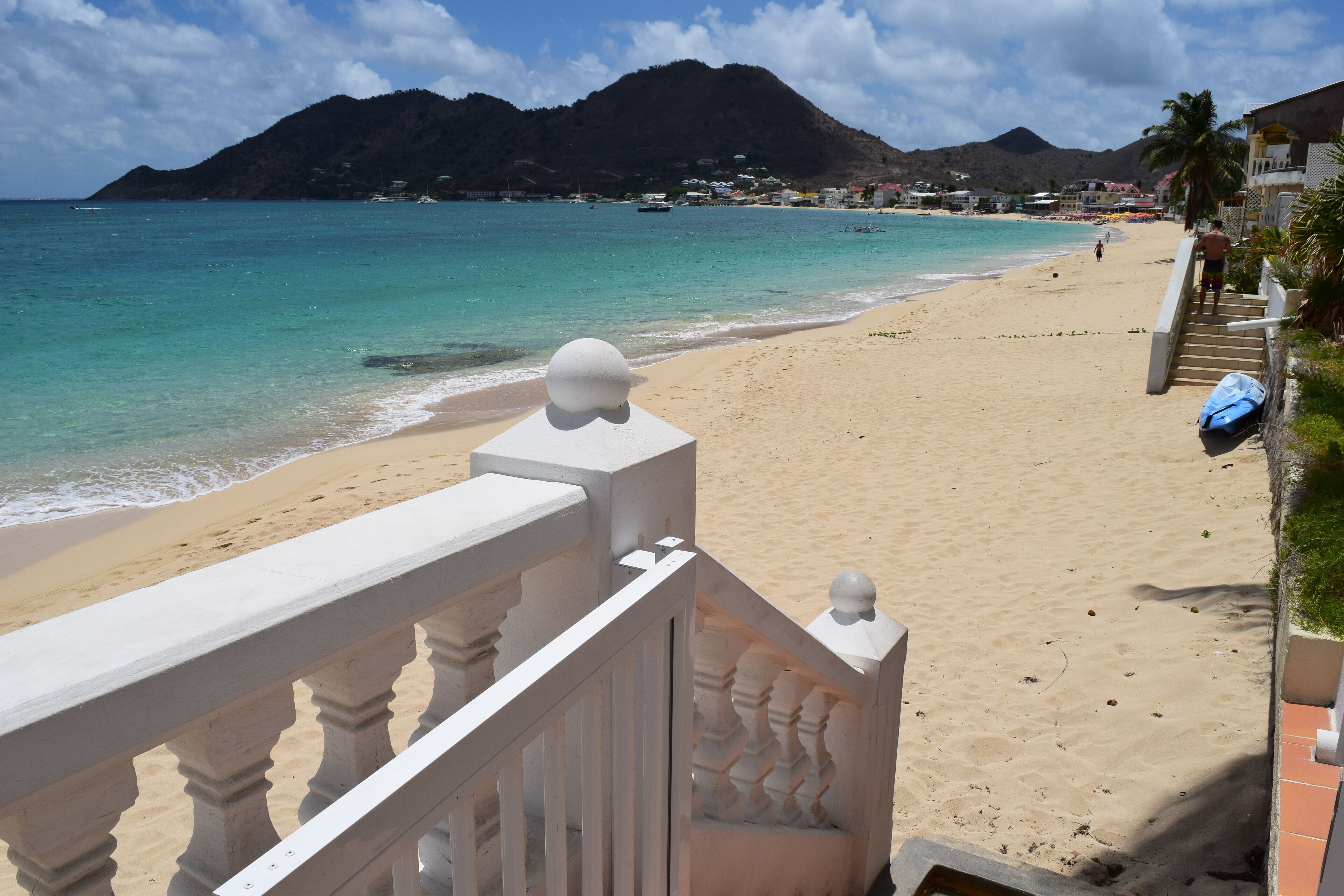 A beach with a white balustrade, clear turquoise water, and a mountain in the background.