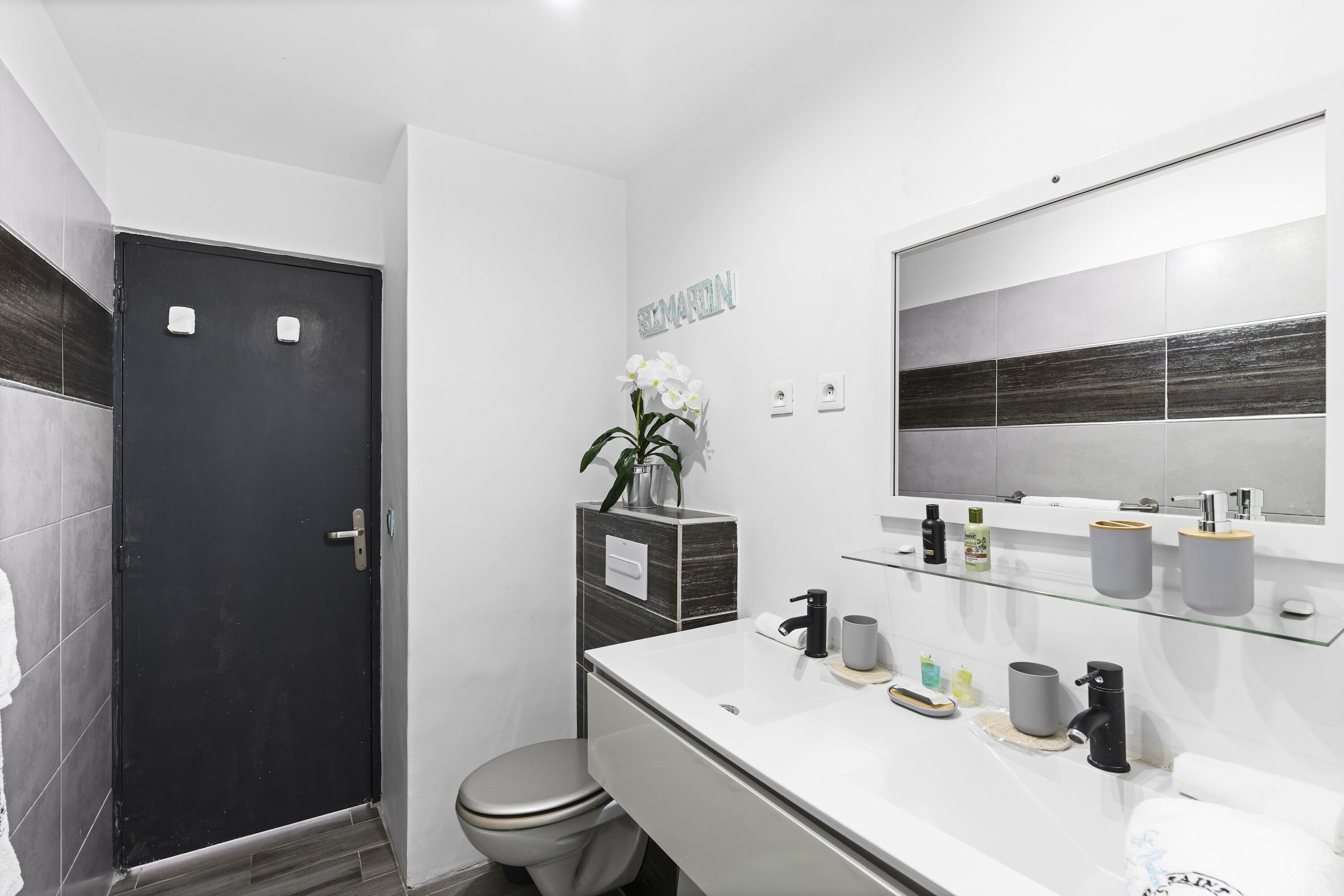 Une salle de bain moderne avec une porte noire, des toilettes, un lavabo avec un miroir et une étagère contenant des articles de toilette.