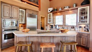 A spacious kitchen with a large island, wooden stools, stainless steel appliances, and a high ceiling with exposed beams. - Stonegate #14 Luxury Cabin with Hot Tub - Walking Distance to Village & Gondola! (Mammoth Lakes)