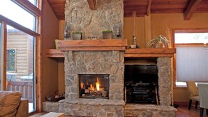A stone fireplace with a TV and entertainment system, wooden beams, and a view of the outdoors. - Stonegate #14 Luxury Cabin with Hot Tub - Walking Distance to Village & Gondola! (Mammoth Lakes)