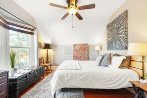 A bedroom with a large bed, a ceiling fan, a brick accent wall, and a window with a view of trees.