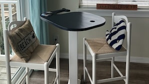 A room with a round table, two chairs with beach-themed cushions, a hanging light fixture, and a window with blinds.