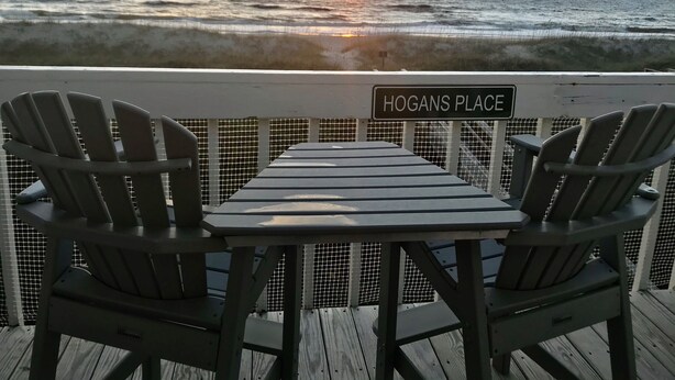 A deck with two Adirondack chairs and a table, overlooking the ocean with a sunset. A sign reads "HOGANS PLACE".
