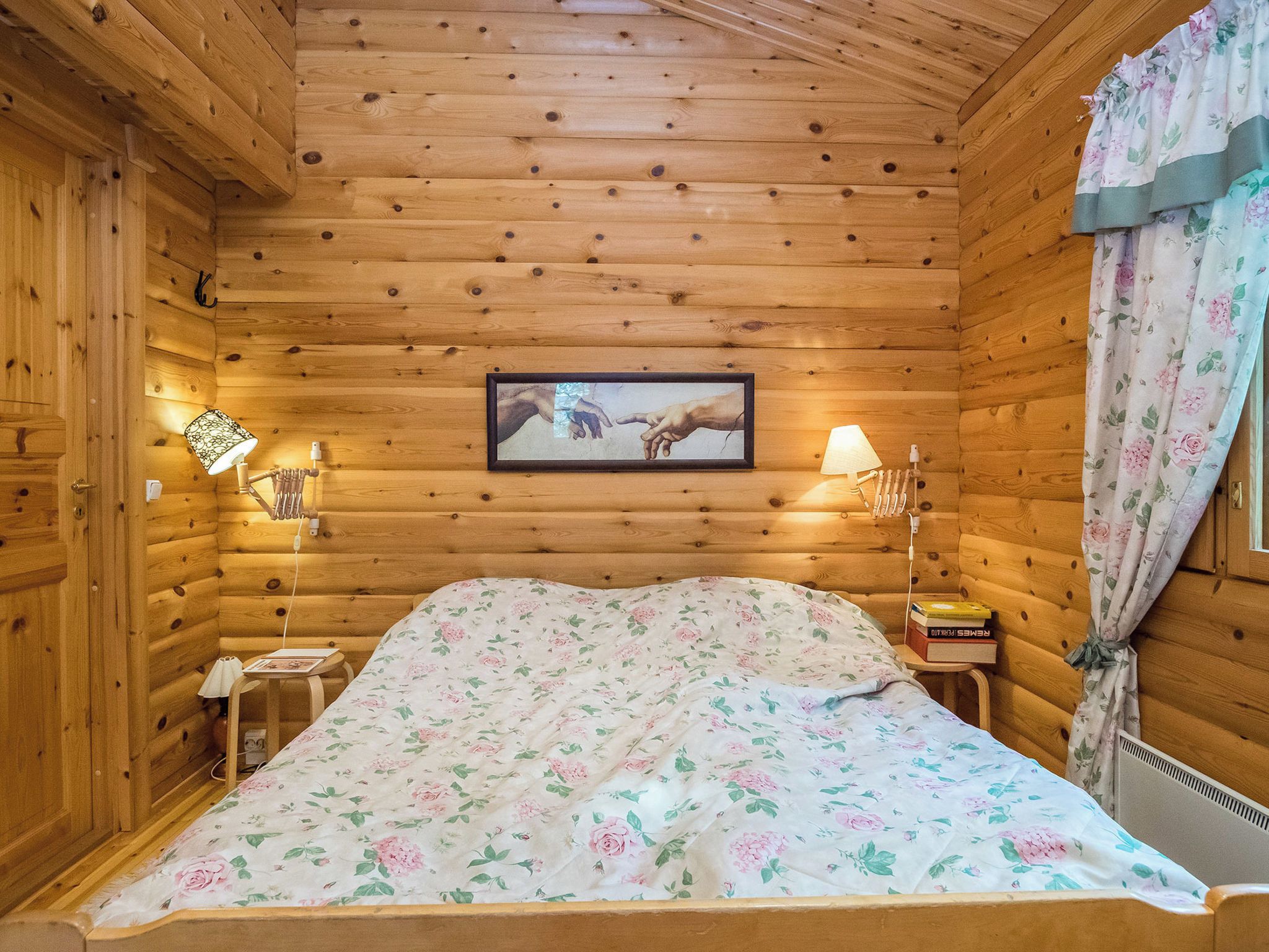 A wooden cabin room with a bed, two bedside lamps, a framed picture on the wall, and a window with curtains.