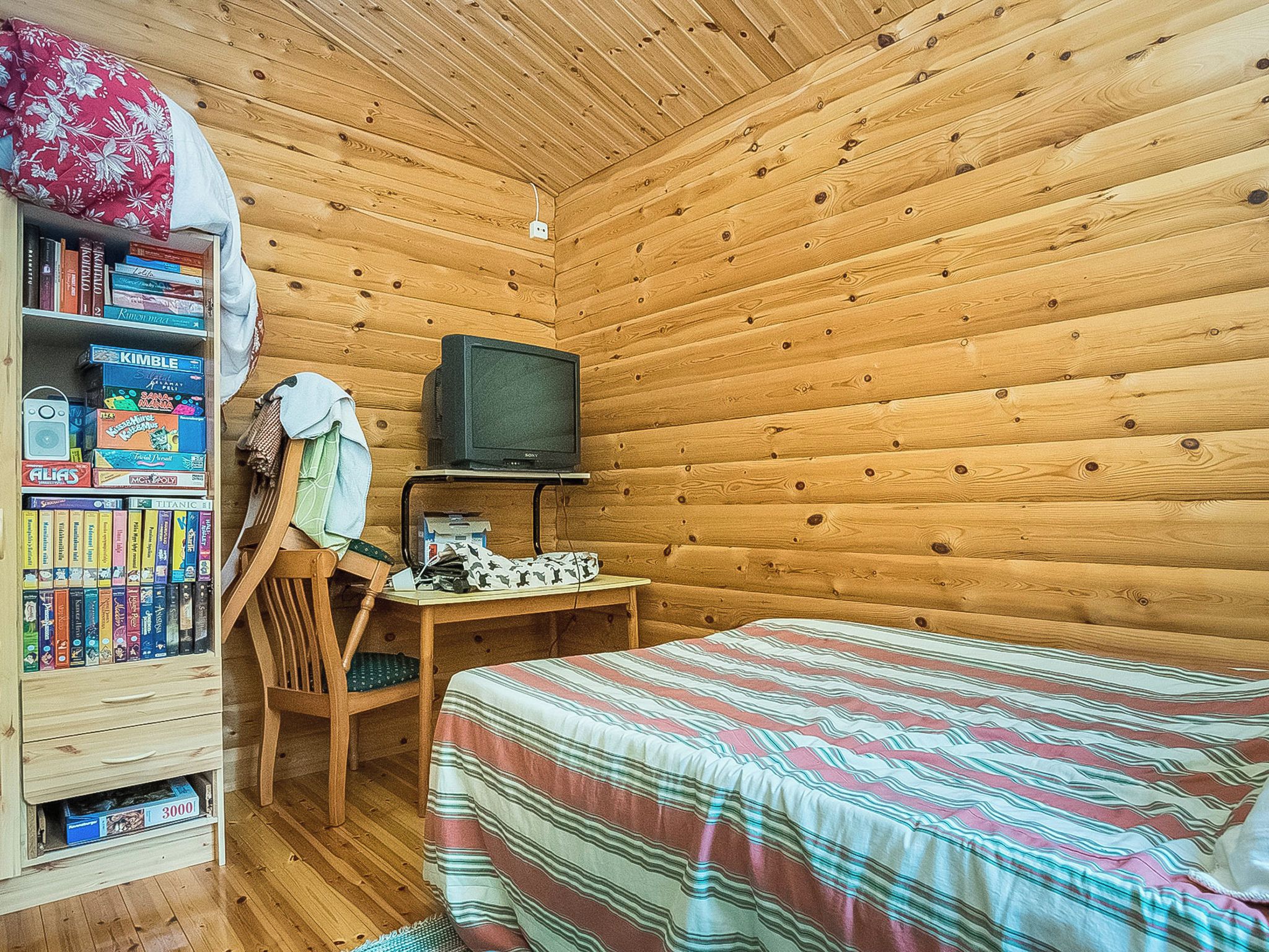 A cozy cabin room with a wooden interior, a bookshelf, a television, a desk, and a bed.