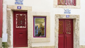 A building with two red doors, each with a unique decorative pattern. There are two visible windows, one with a purple frame and another with a yellow frame. The building has a textured facade and numbered plaques above the doors. - Portuguese Soul Alfama | Terrace & River View | Sleeps 6 (Lisboa)