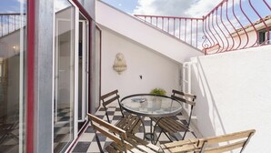 A glass table with chairs, a potted plant, and a wall-mounted light fixture in a courtyard with a checkered floor. - Portuguese Soul Alfama | Terrace & River View | Sleeps 6 (Lisboa)