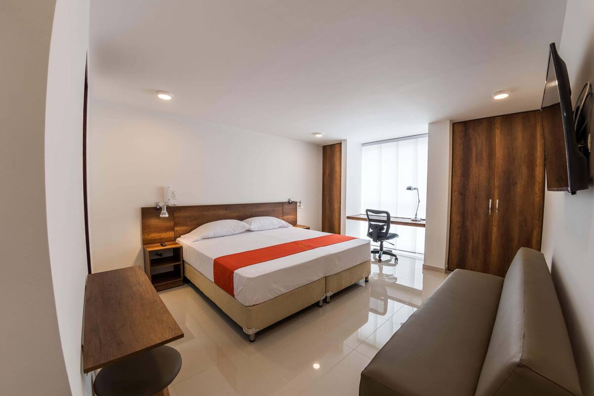 A modern hotel room with a bed, a desk, a chair, a sofa, and a TV mounted on the wall.