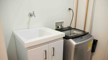 Un baño pequeño con un lavamanos blanco, una lavadora de acero inoxidable y un enchufe eléctrico en la pared.