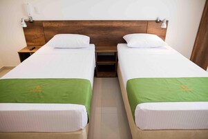 A hotel room with two single beds, each with a green bedspread and white pillows. There is a wooden headboard, a nightstand with a lamp, and a small shelf between the beds.
