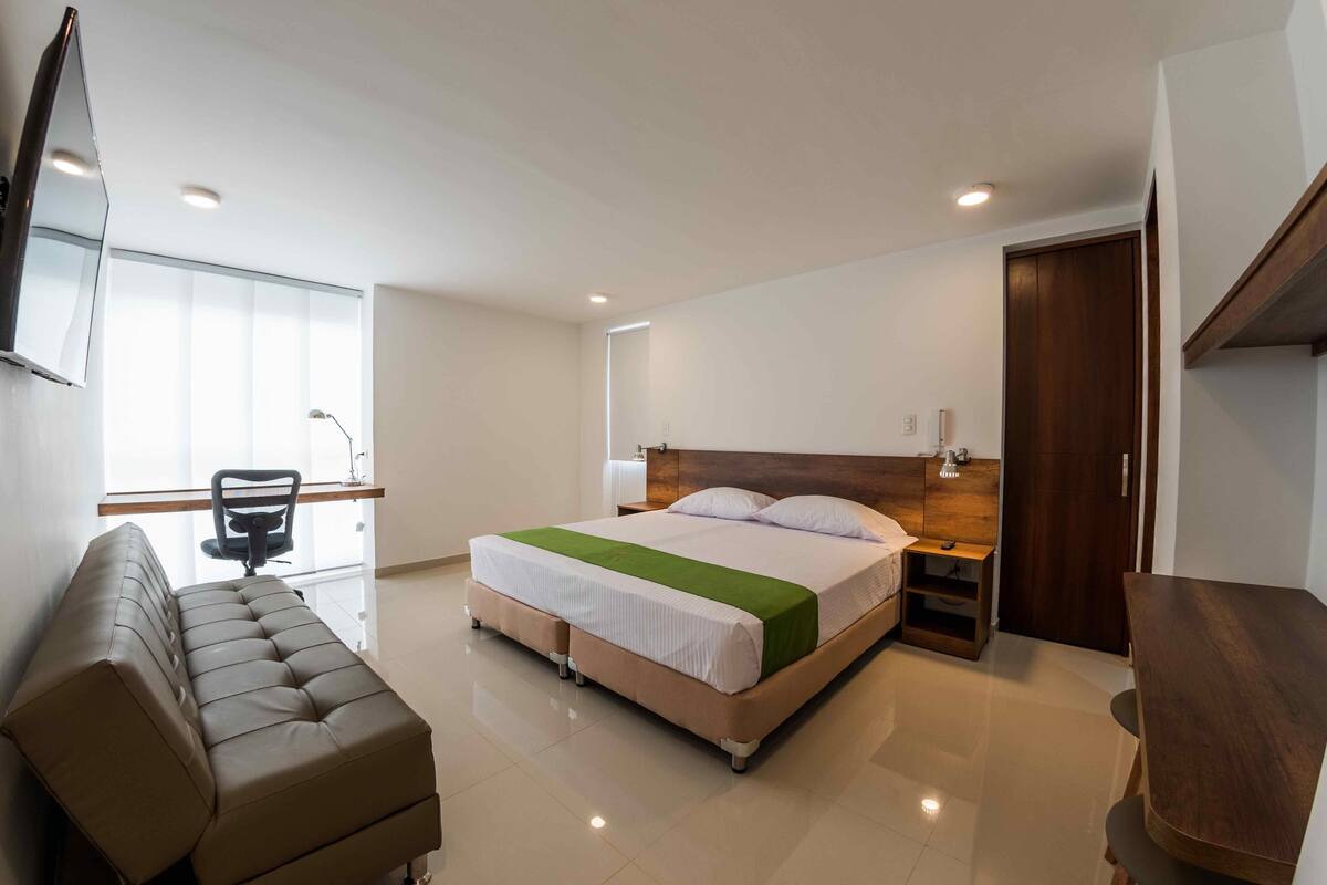 A modern hotel room with a large bed, a sofa, a desk, and a TV mounted on the wall.