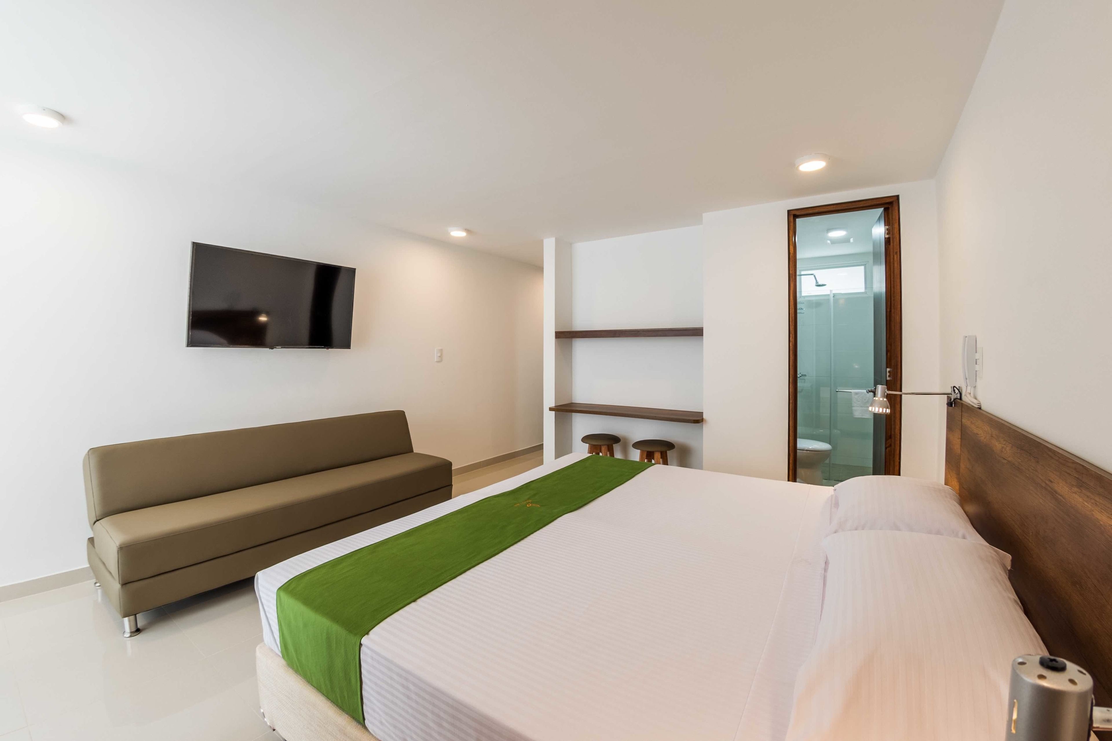 A modern hotel room with a bed, a sofa, a television, and a bathroom visible through a glass door.