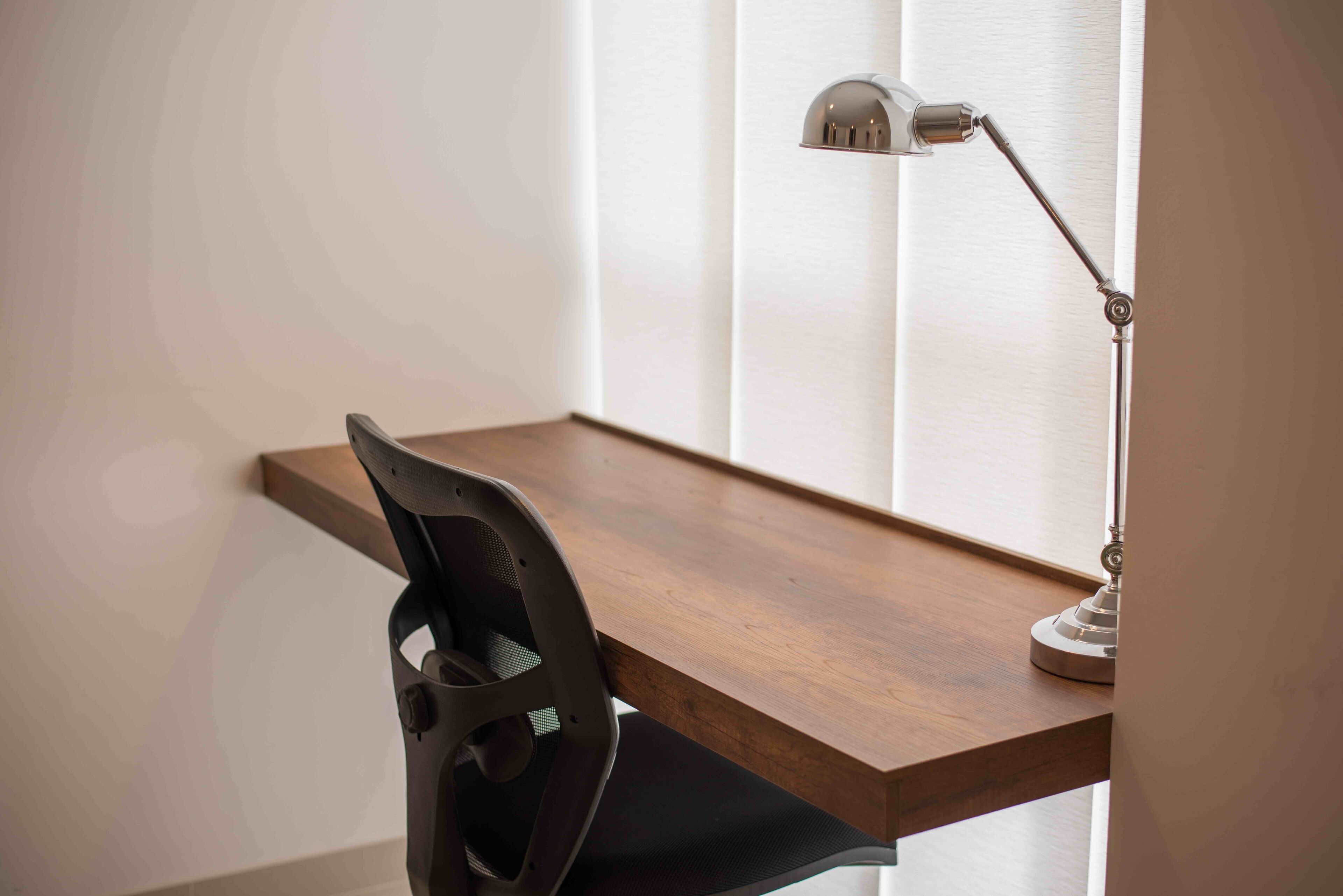A desk with a chair, a lamp, and a window with blinds.