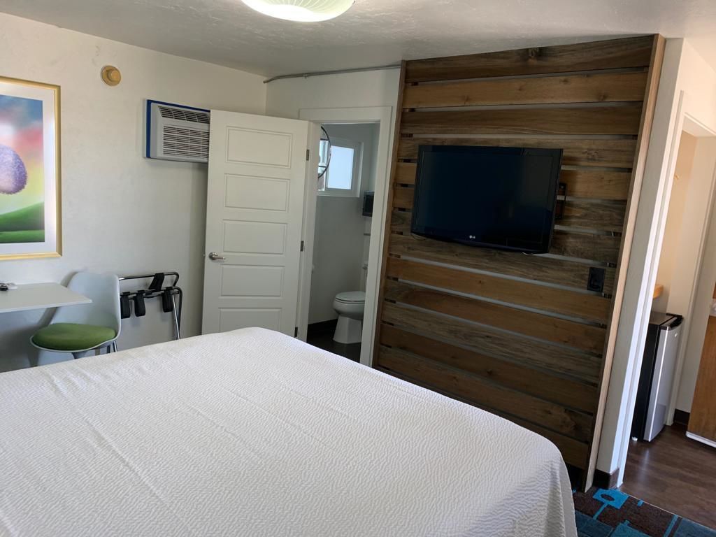 Una habitación de hotel con cama, televisor montado en una pared de madera, puerta que da a un baño y una pequeña mesa con una silla.