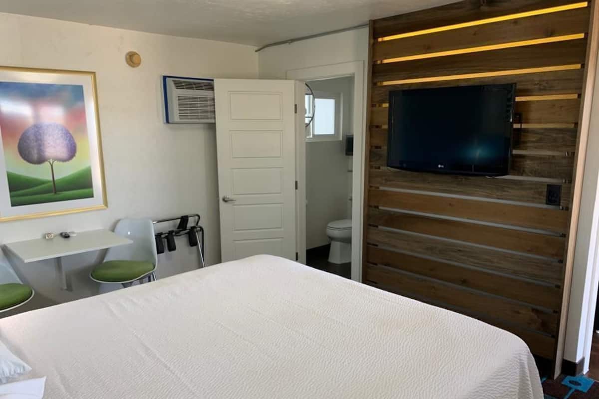 A hotel room with a bed, a desk with a chair, a television mounted on a wooden wall, and a door leading to a bathroom.
