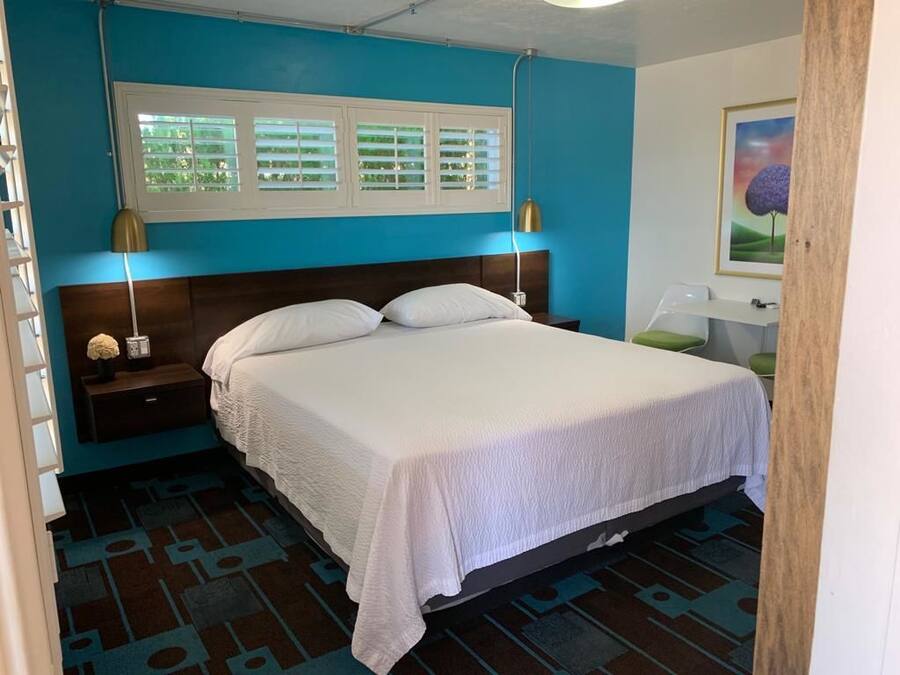 Quarto com cama, mesas de cabeceira, janela com persianas, luminária e um quadro na parede.