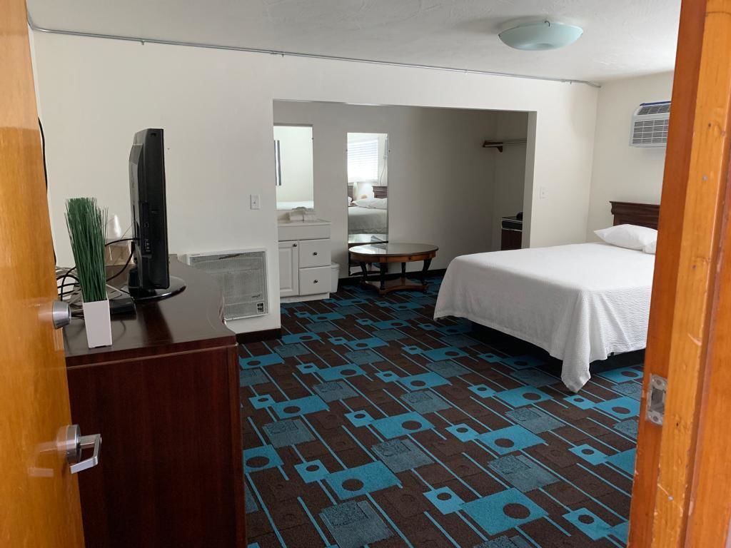 A hotel room with a bed, a desk with a television, a mirror, a bathroom visible through an archway, and a patterned carpet.