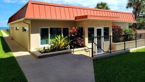 A single-story building with a red roof, a concrete walkway, and a small garden area with red flowers. - 111 - Poolside 1 bedroom closest to the water (New Smyrna Beach)