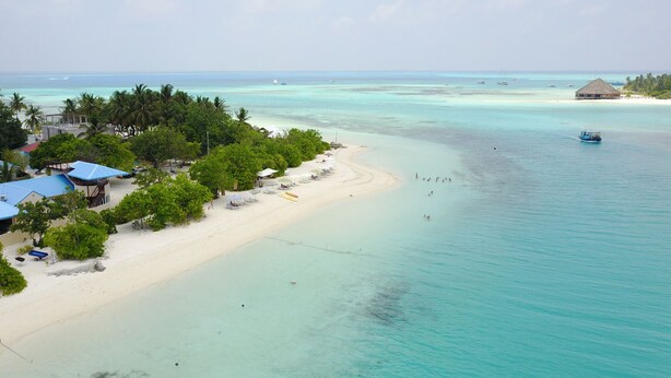 A beach resort with a sandy shoreline, clear turquoise waters, and a few boats. There are structures with blue roofs and umbrellas along the beach. - Infinity Dhiffushi (Dhiffushi)