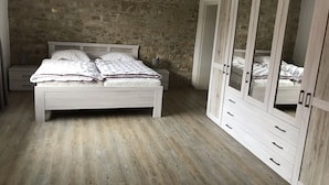 A bedroom with a stone wall, a wooden floor, a white bed with a quilt, and a large wardrobe with mirrored doors.