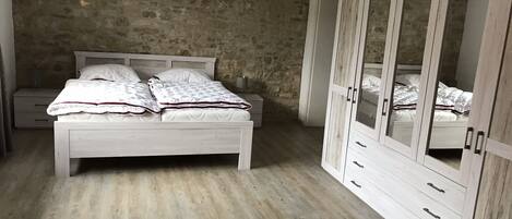 Un dormitorio con pared de piedra, suelo de madera, cama blanca con colcha y un gran armario con puertas de espejo.