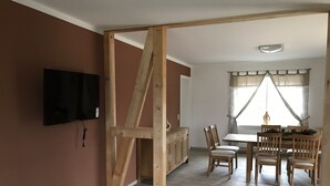 A room with a wooden structure, a TV mounted on the wall, a dining table with chairs, and a window with curtains.