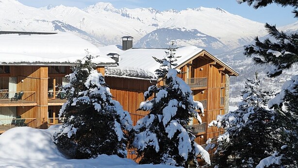 Un chalet in legno con diversi balconi e un tetto innevato, circondato da alberi imbiancati dalla neve e montagne sullo sfondo.
