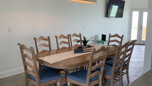 A dining area with a wooden table and chairs, a hanging light fixture, a flat-screen TV mounted on the wall, and a door leading to an outdoor area. - Sunset Cove-4 BR, 2 1/2 BA Waterfront Home. Sleeps 12-Private pier! (Rockport)